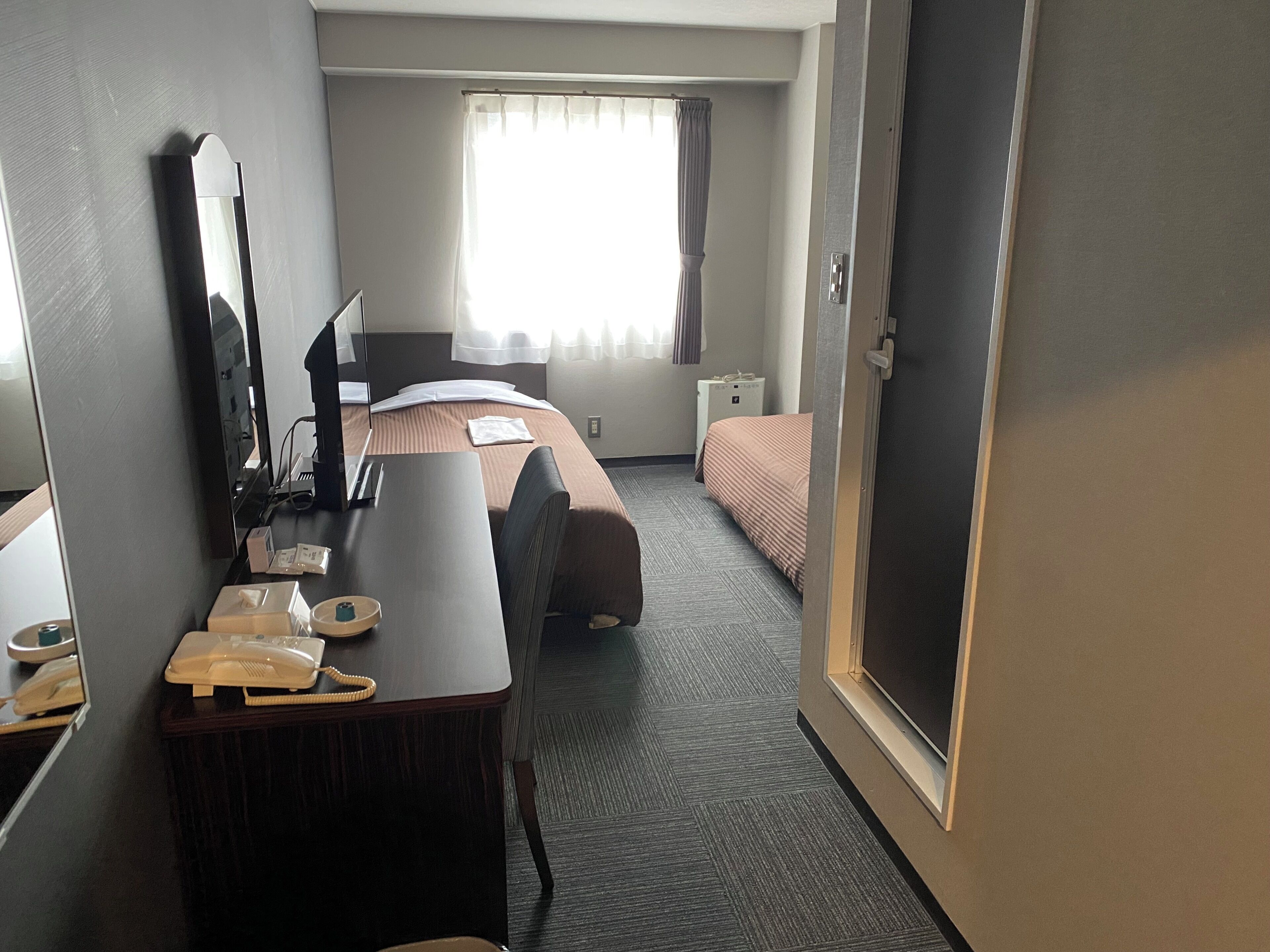 A hotel room with a bed, a desk with a telephone, a mirror, and a window with curtains.