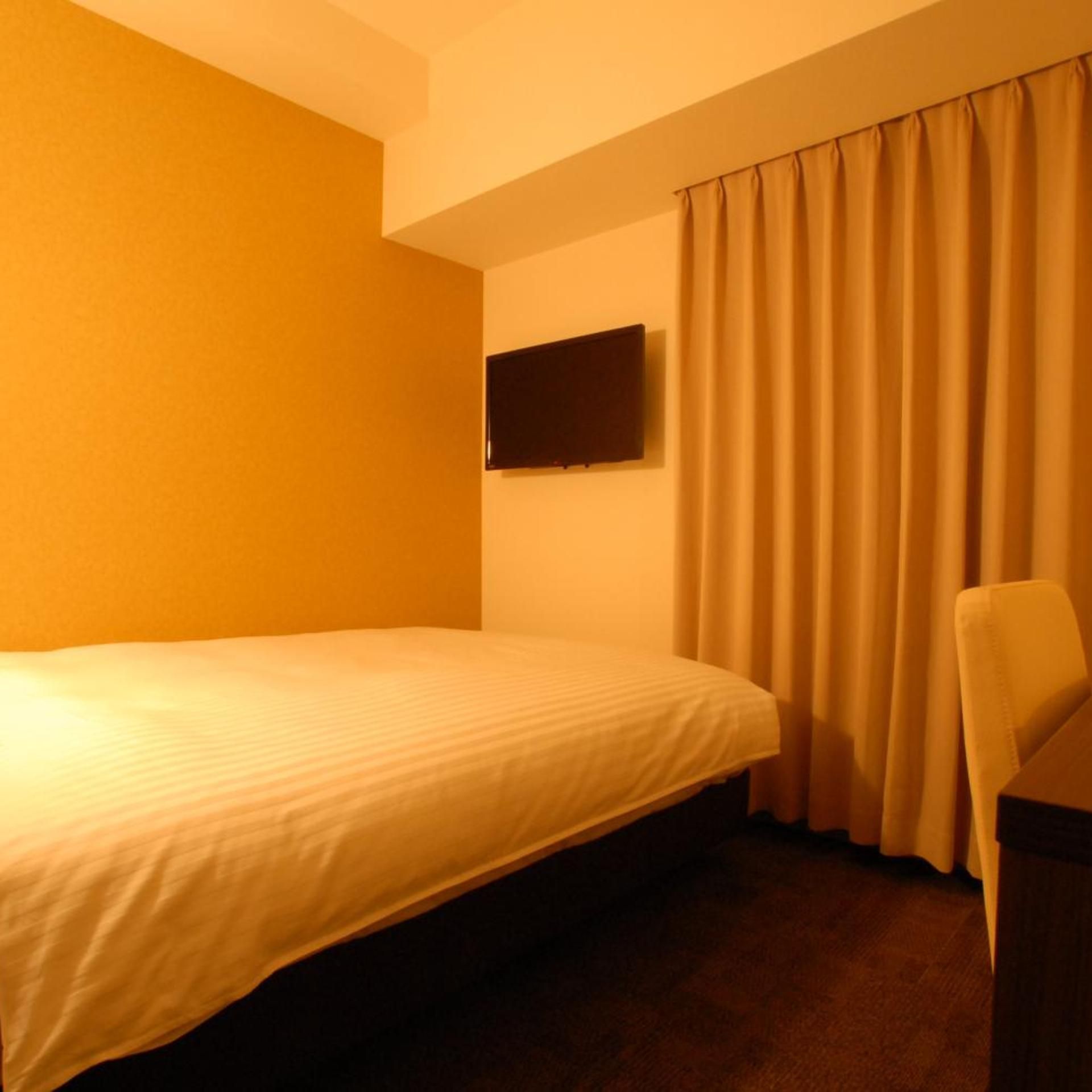 Une chambre d’hôtel avec un lit simple, un téléviseur à écran plat fixé au mur et une chaise.