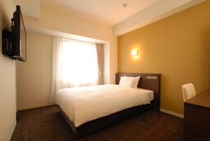 Habitación de hotel con una cama, un escritorio, una silla, un televisor montado en la pared y una ventana con cortinas.