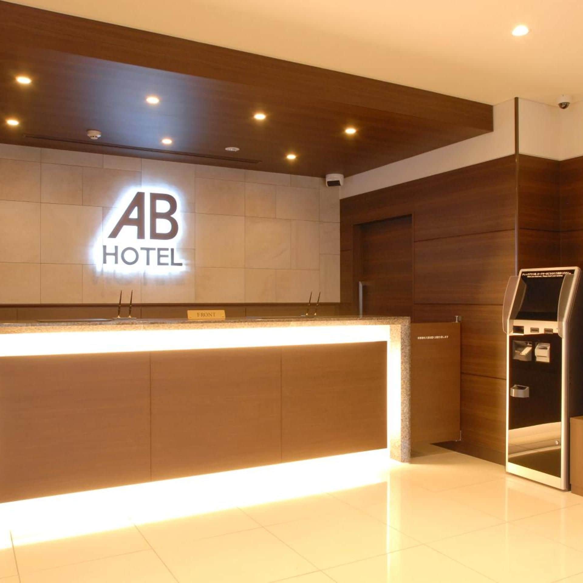 Un comptoir d'accueil d'hôtel avec une enseigne indiquant « AB HOTEL » et un kiosque numérique situé à droite.
