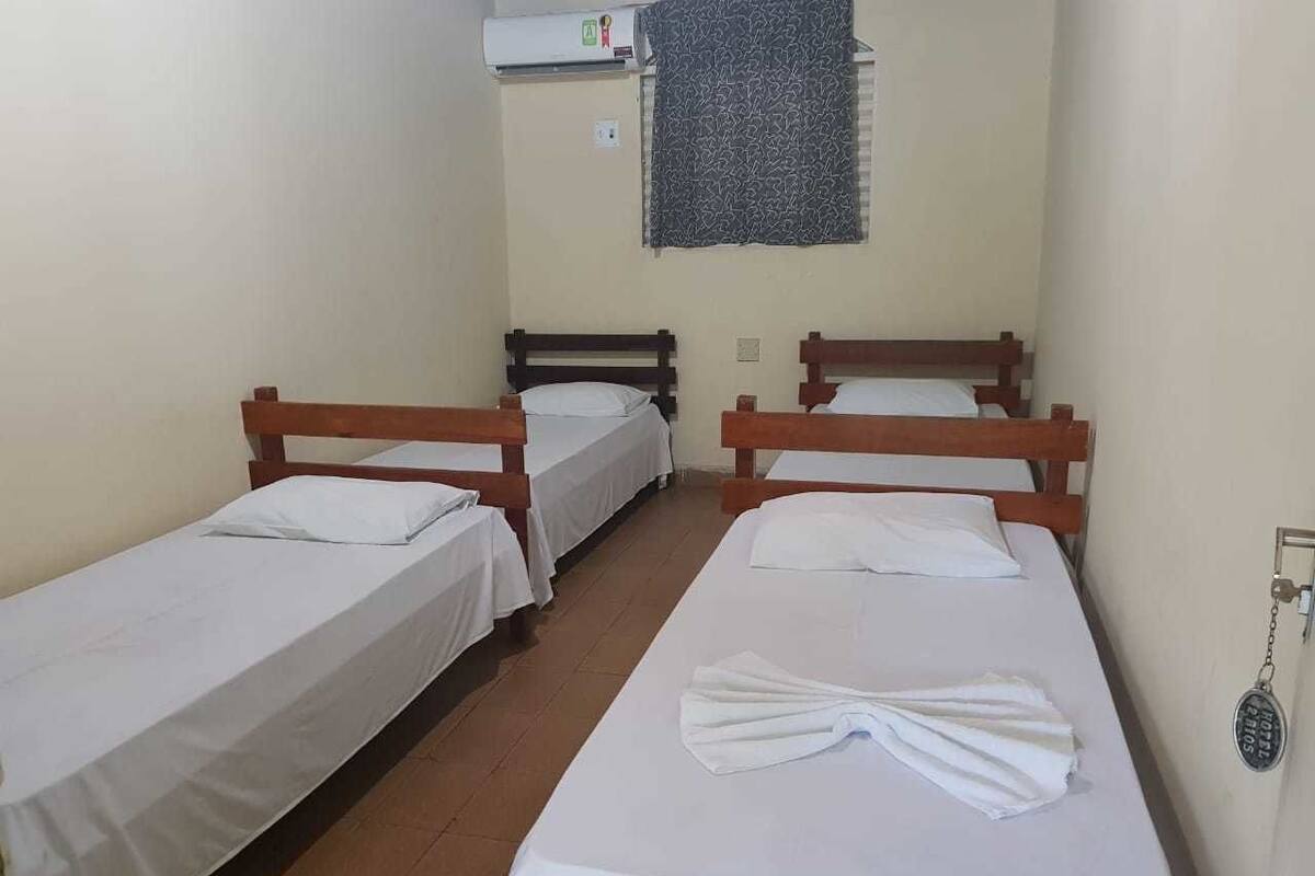A room with three single beds, each with wooden frames and white linens. There is a wall-mounted air conditioner above the beds and a window with a patterned curtain.