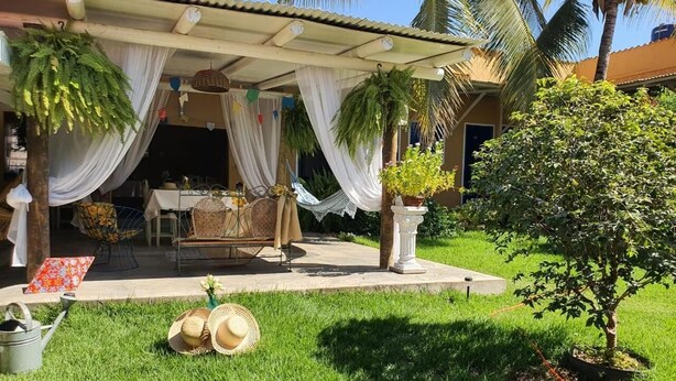 A covered outdoor seating area with a table and chairs, surrounded by lush greenery and palm trees.