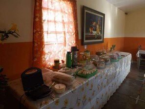 A dining area with a long table covered in a floral tablecloth, various food items, and a laptop on the table.