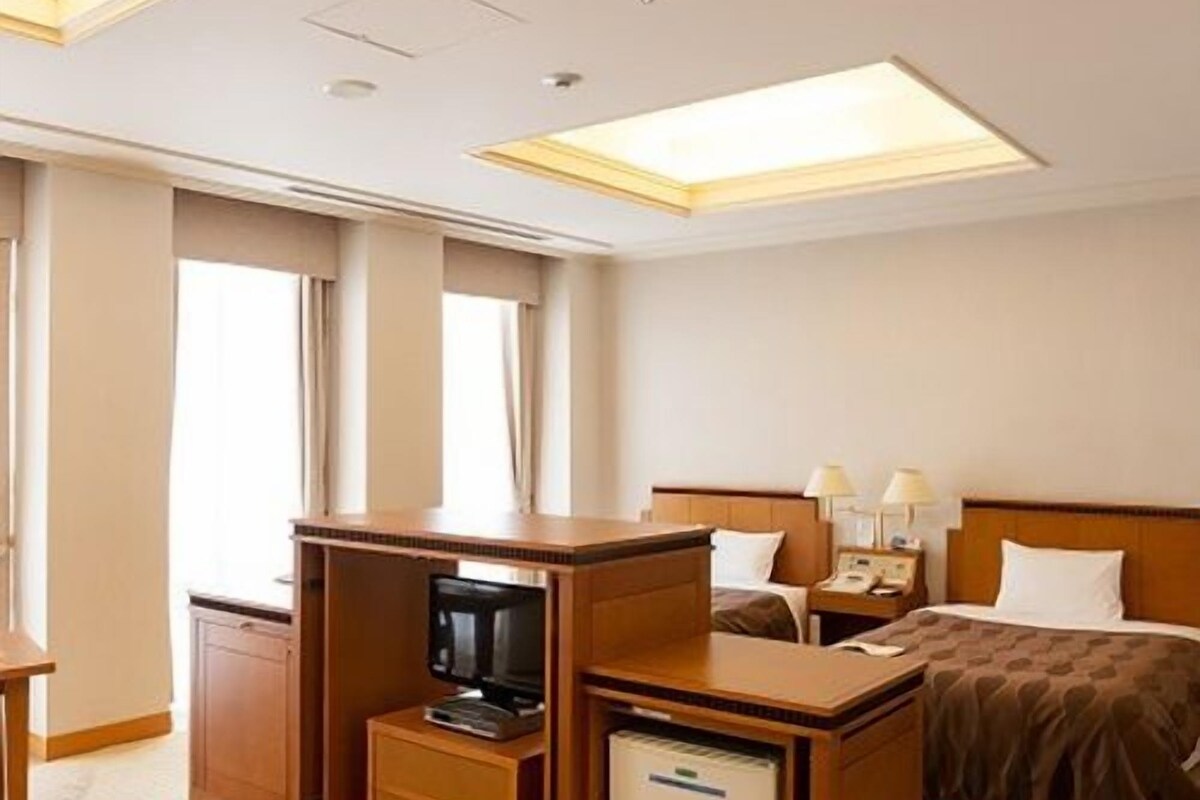 A hotel room with a bed, bedside tables, a desk with a television, and a minibar.