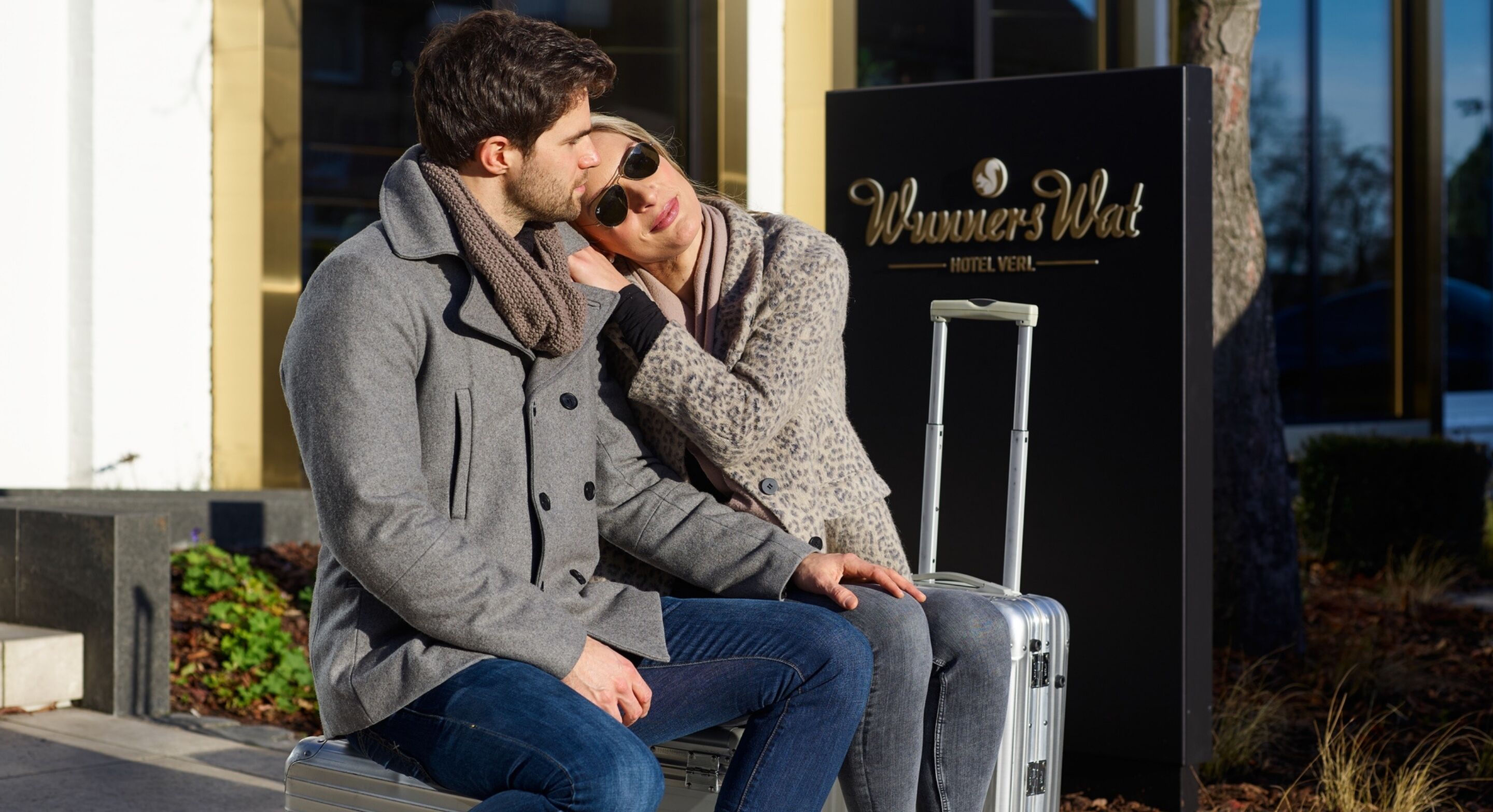 Two people sitting on a bench next to a suitcase with a sign for 'Winners Wat Hotel' in the background.