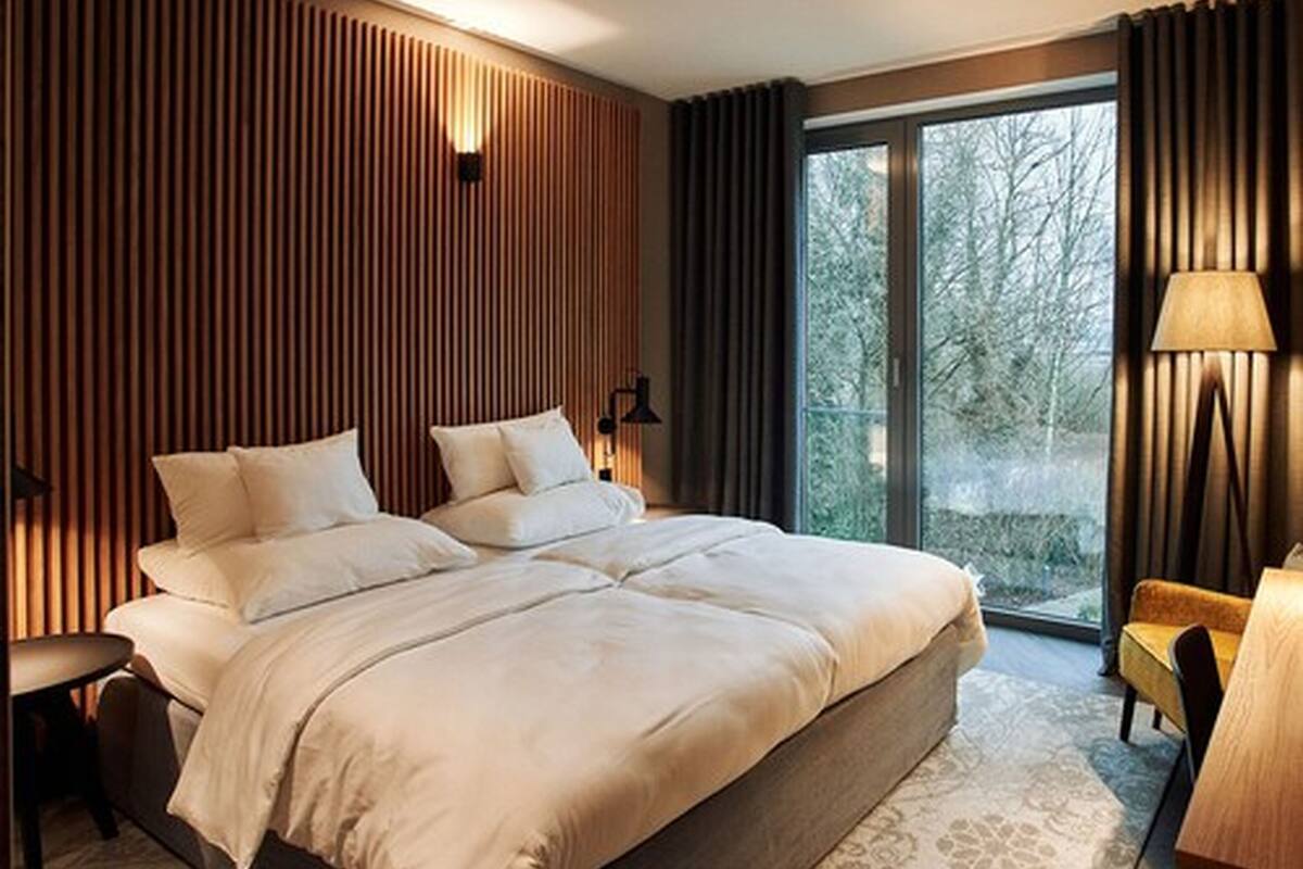 A modern bedroom with a large bed, a bedside table, a lamp, and a view of trees through a large window.