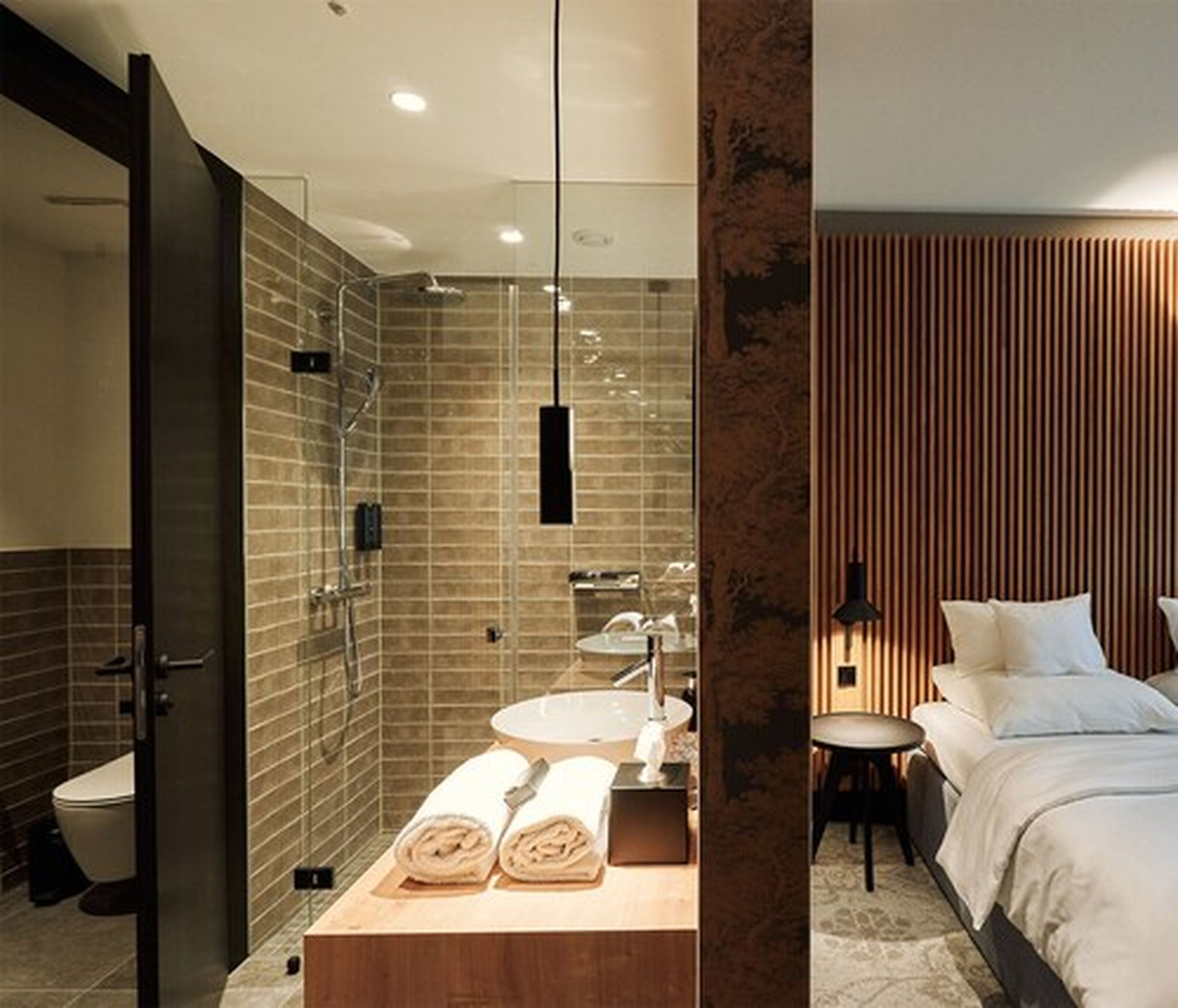 Un baño moderno con cabina de ducha acristalada, tocador de madera con lavamanos y una cama bien hecha con cabecero de madera.