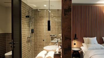 Un baño moderno con cabina de ducha acristalada, tocador de madera con lavamanos y una cama bien hecha con cabecero de madera.