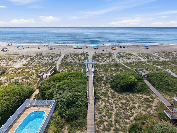 A beach with a wooden boardwalk leading to a pool area, surrounded by dunes and beach umbrellas.