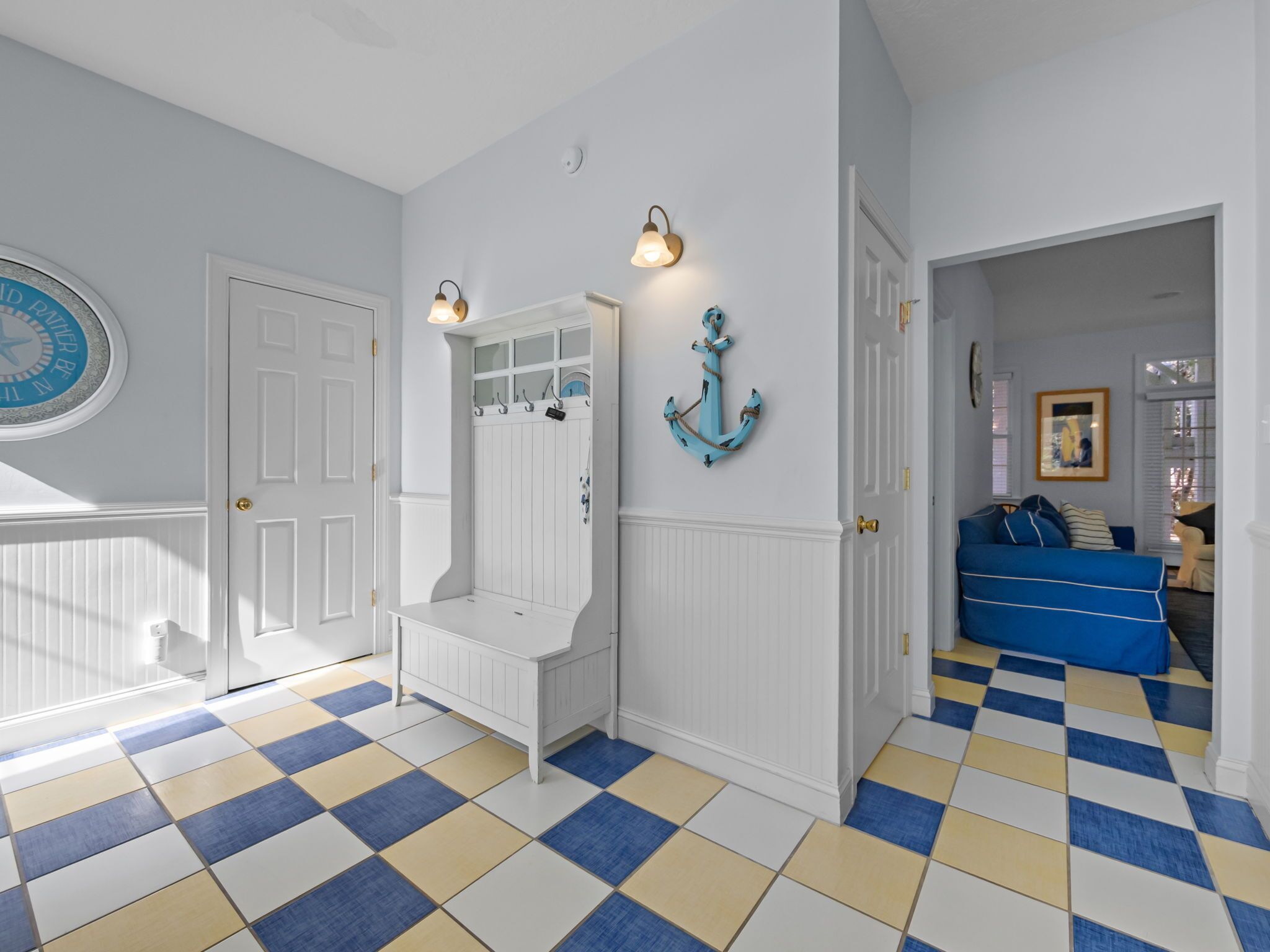 Un couloir avec un sol à carreaux, un banc blanc, un porte-manteau fixé au mur et une porte ornée d’un miroir rond.