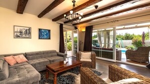 A living room with a grey sofa, a wooden coffee table, wicker chairs, and a large window with a view of a swimming pool and greenery. - Spacious holiday house / pool / jacuzzi / playground / 6+2 (Rakalj)