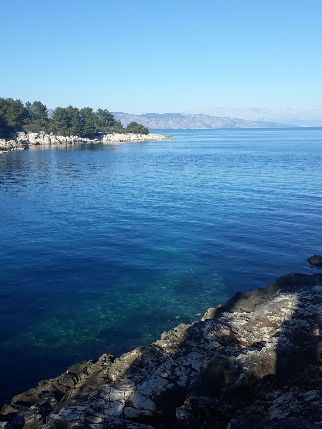 Eine Küstenlandschaft mit klarem, blauem Wasser, felsiger Uferlinie und Bergen im Hintergrund.
