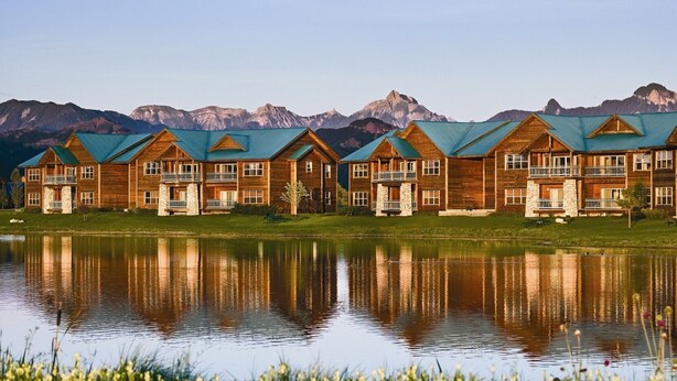 A row of wooden cabins with green roofs, a reflecting lake, and mountains in the background.
