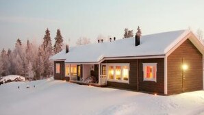 A wooden cabin with a snow-covered roof, a porch, and a lit interior.