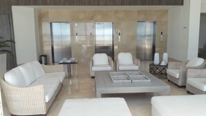 A modern living room with a beige tiled wall, two white sofas, a coffee table, and two chairs. There are two elevators in the background. - Palmetto Sunset (Cartagena)