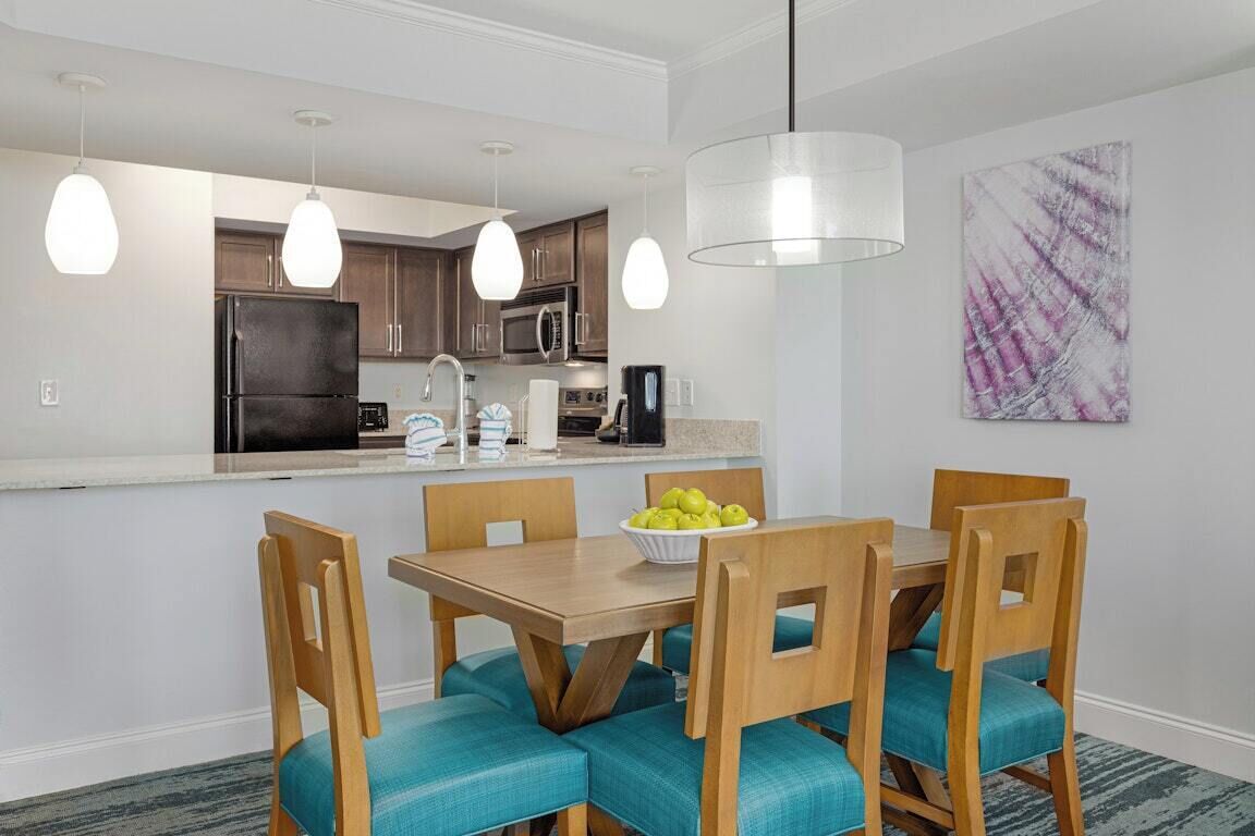 A modern kitchen with a central island, wooden chairs with teal cushions, and pendant lighting.