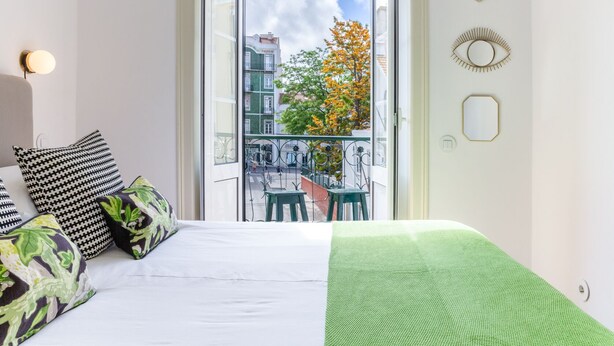 Ein Schlafzimmer mit einem Bett, einer grünen Decke, einem schwarz-weißen Kissen und Blick auf einen Balkon mit Geländer und einem Baum.