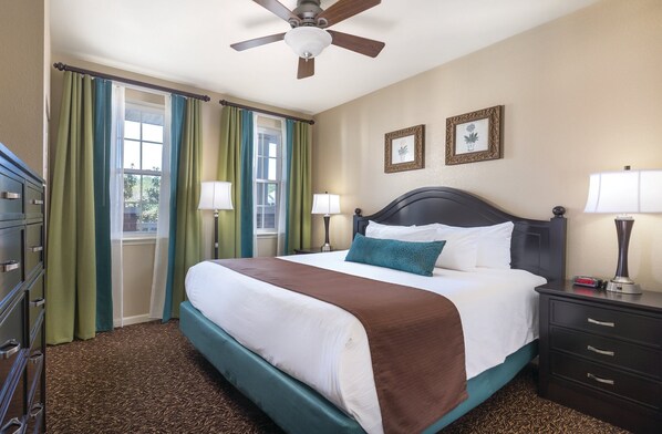 Habitación de hotel con cama, dos lámparas de noche, ventilador de techo, cómoda y ventana con cortinas.