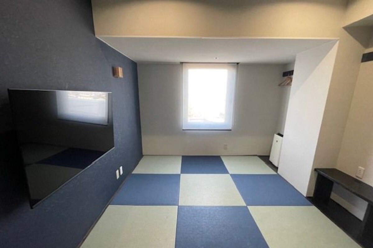 Une pièce avec un sol à carreaux, un téléviseur à écran plat fixé au mur, une fenêtre avec des stores et un petit meuble.