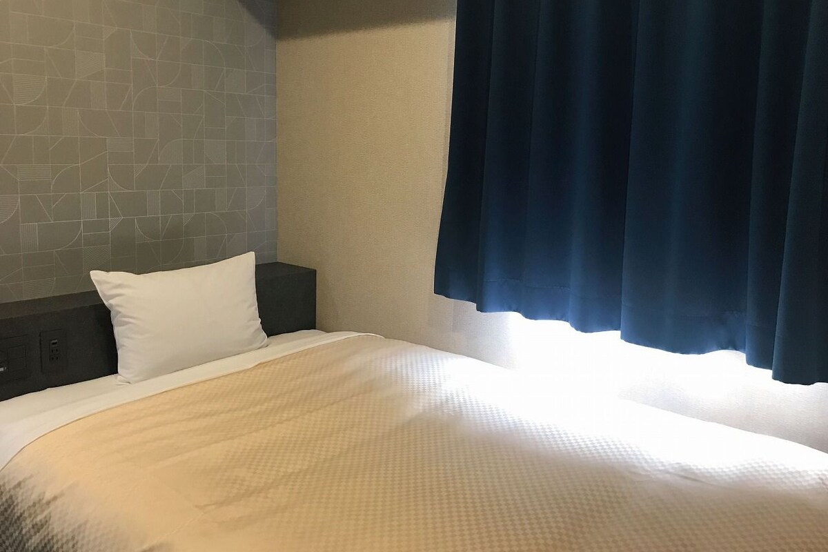 Habitación de hotel con una cama grande, cabecera oscura, almohada blanca y ventana con cortinas oscuras.
