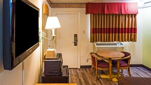 Et kompakt hotelværelse med fjernsyn, et skrivebord med lampe, et lille bord med to stole, et vindue med stribede gardiner og en fastgjort aircondition.