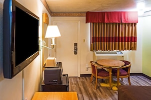 Habitación de hotel compacta con televisión, escritorio con lámpara, pequeña mesa con dos sillas, ventana con cortinas a rayas y aire acondicionado instalado en la pared.