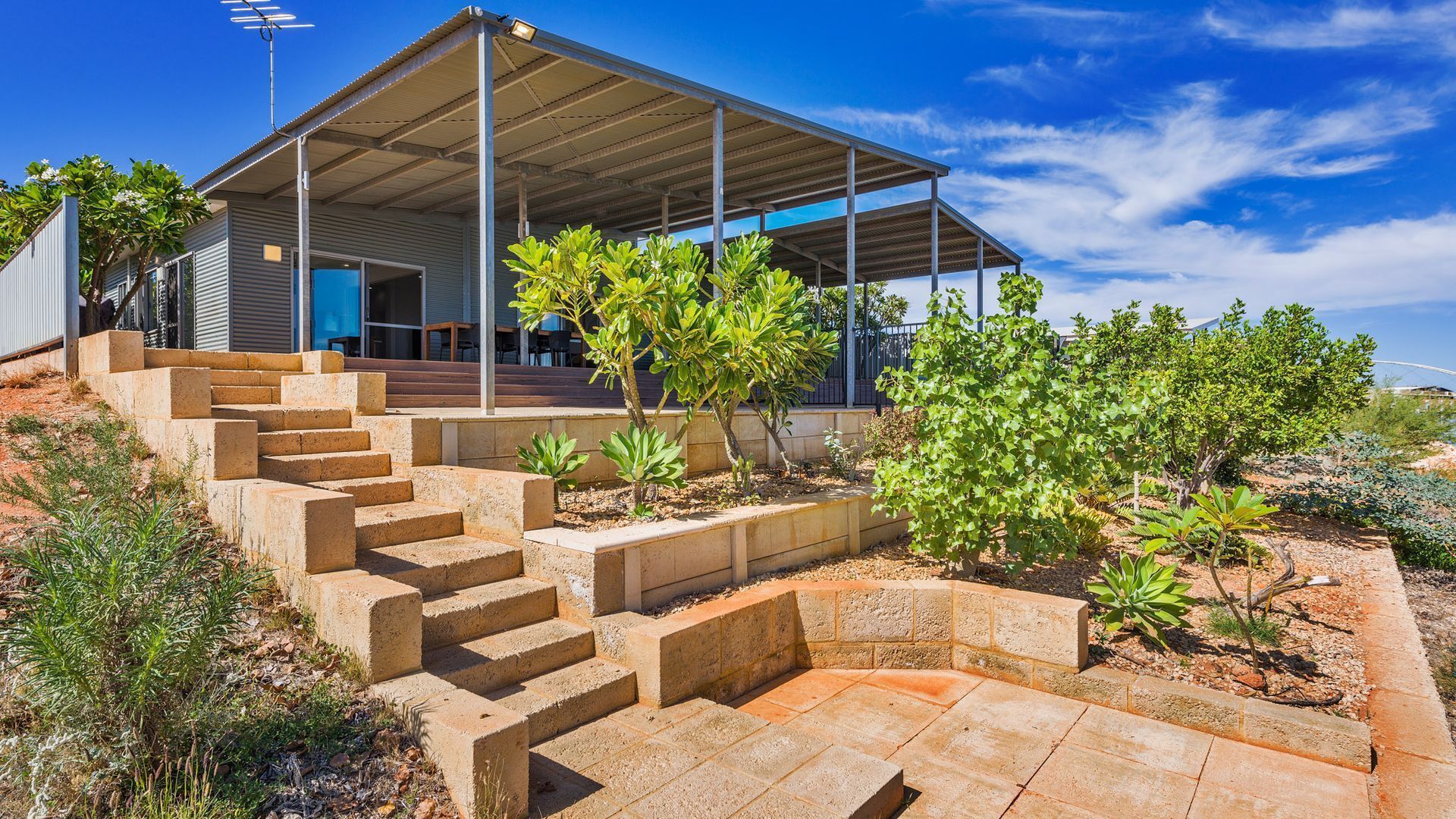 A modern house with a covered outdoor area, concrete steps leading to a door, and various plants.