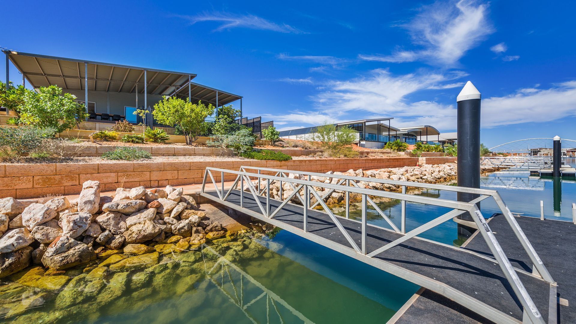 A modern building with a covered outdoor area, a bridge over a water feature, and a marina in the background.