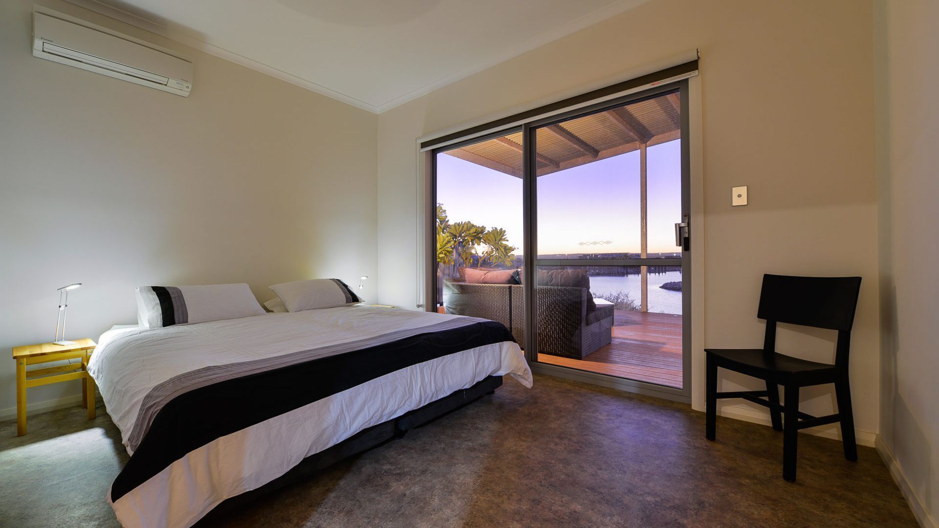 A bedroom with a bed, a chair, a nightstand, and a view of a balcony with a table and chairs.