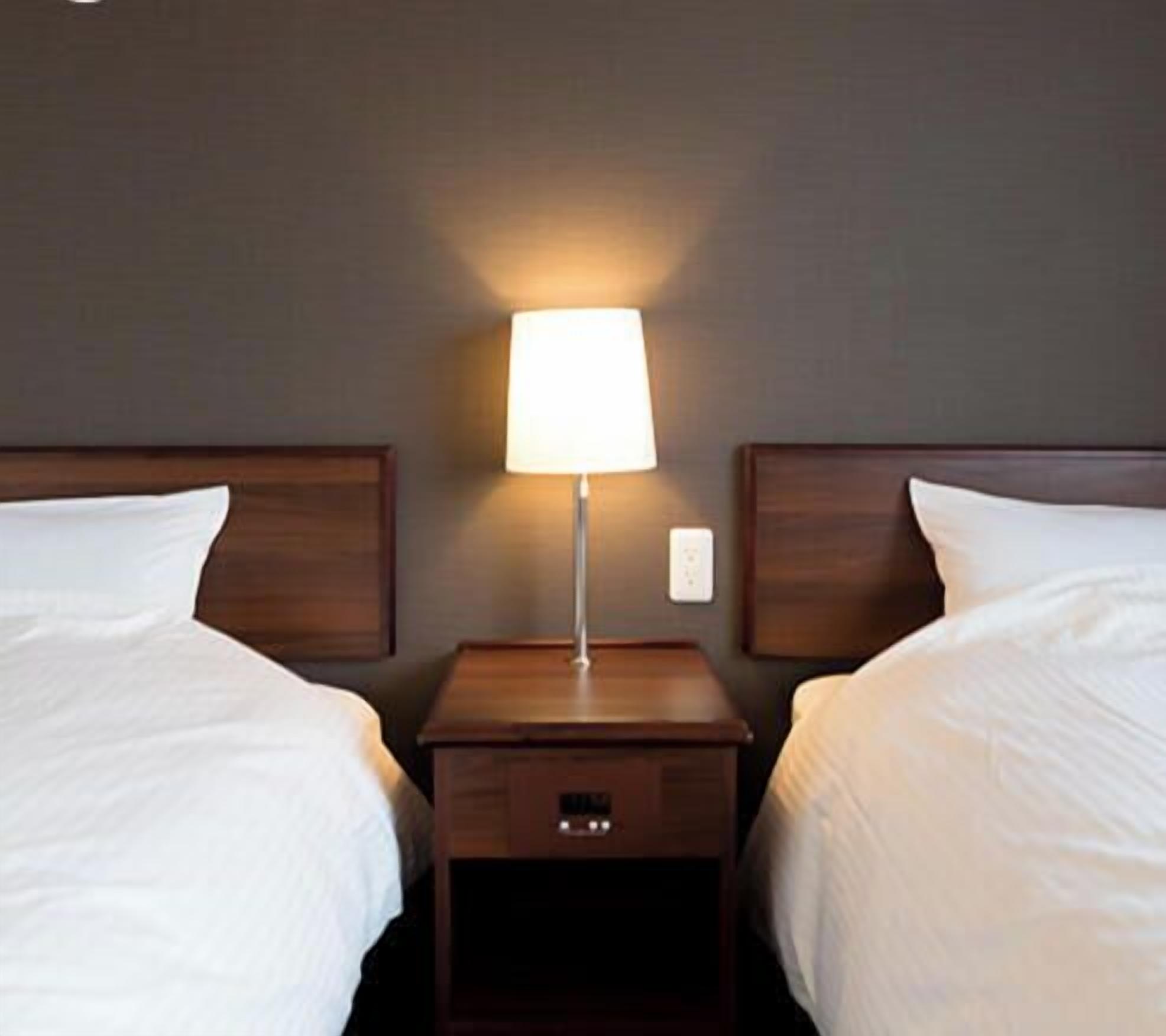 Hotellihuone, jossa on kaksi erillistä sänkyä, yöpöytä lampun kanssa ja pieni laatikosto.