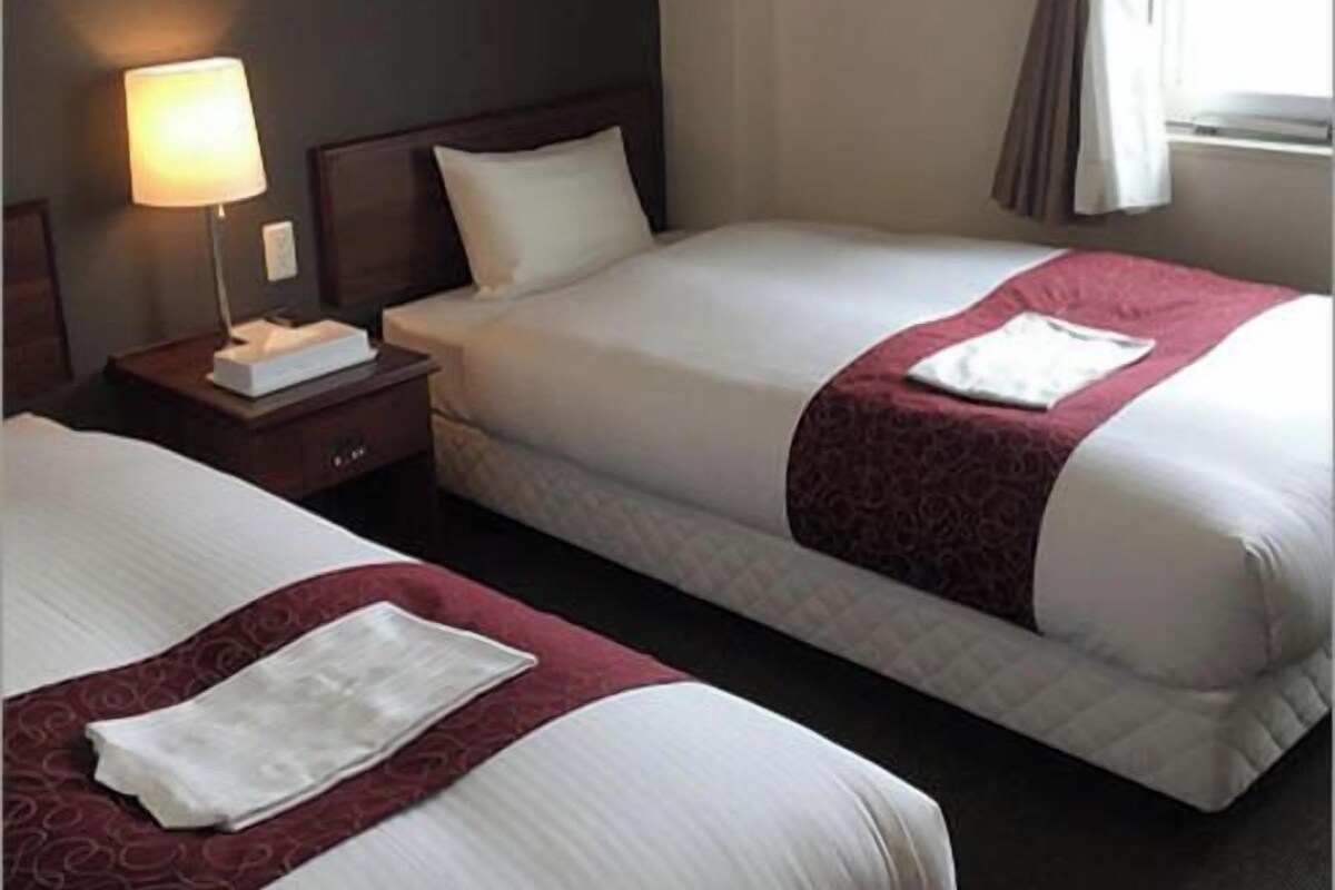 Una habitación de hotel con dos camas, una mesita de noche con lámpara y un ventanal con cortinas.