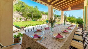 Un área de comedor al aire libre cubierta con una mesa dispuesta para seis personas, rodeada de sillas y vistas a un jardín y un muro de piedra.