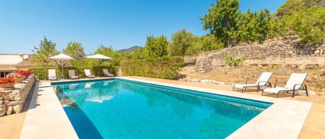 Una piscina rectangular con agua azul cristalina, rodeada por un muro de piedra y un sendero pavimentado. Hay sillas de descanso blancas y una pequeña mesa con un cuenco de fruta a la izquierda.