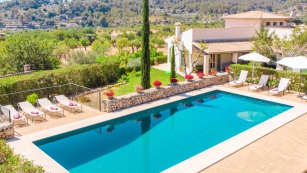 Piscina con sillas de descanso y pared de piedra, rodeada de vegetación y vistas a colinas.