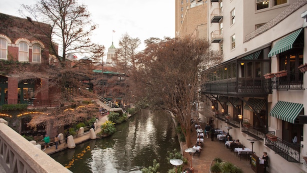 A riverside walkway with tables and chairs, surrounded by buildings and trees.
