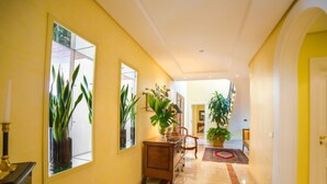 A hallway with yellow walls, large mirrors, potted plants, a wooden cabinet, and a patterned rug.
