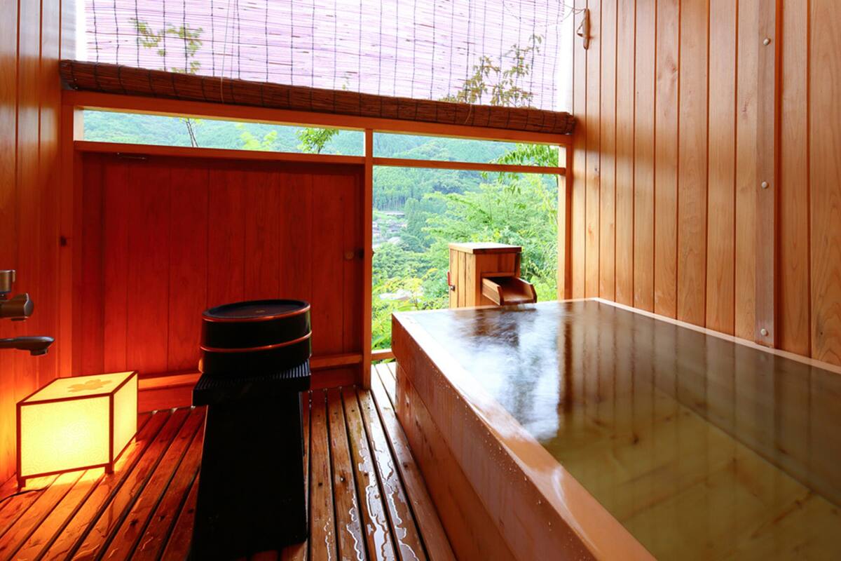 A wooden room with a hot tub, a small wooden stool, and a window with a view of greenery.