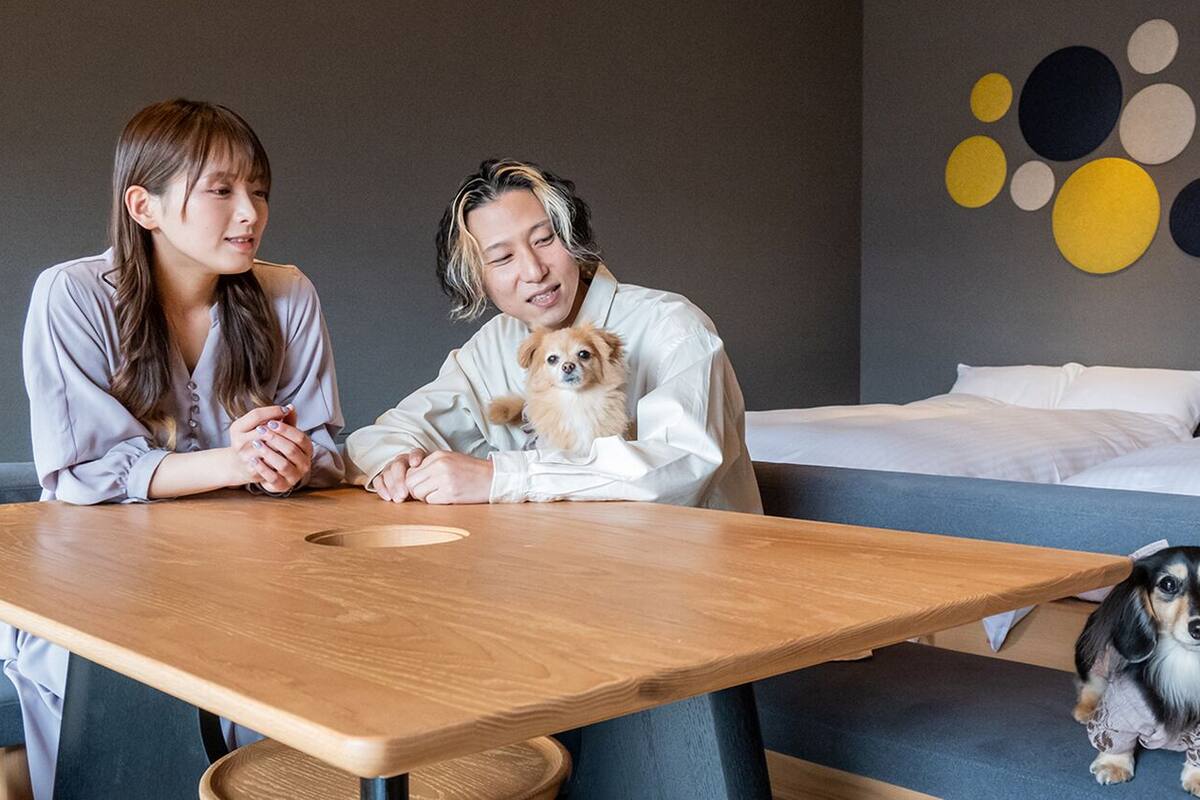 Two people sitting at a wooden table in a room with a bed and a wall with circular patterns.