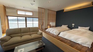 Quarto de hotel moderno com uma janela ampla, um sofá, duas camas e vista para um corpo d'água.