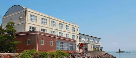 A coastal building with a large windowed section, a smaller section with a flat roof, and a lighthouse in the distance.