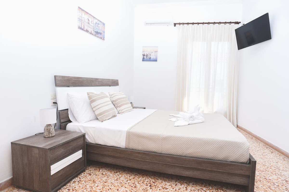 Un dormitorio ordenado con estructura de cama de madera, ropa de cama blanca, una mesita de noche con lámpara y un televisor de pantalla plana montado en la pared.