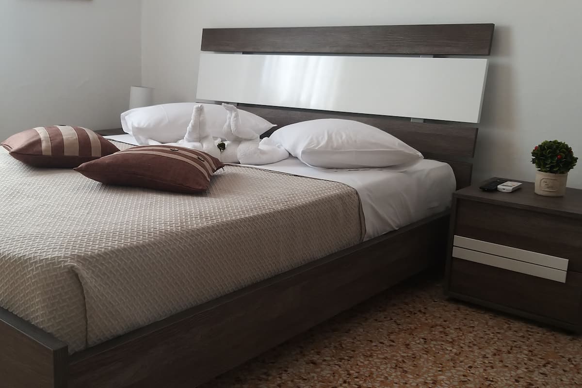 Una cama bien hecha con almohadas blancas y marrones, una manta blanca y una pequeña planta en maceta sobre la mesita de noche.