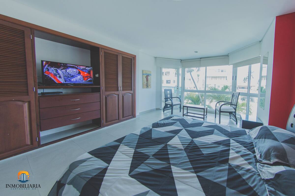 A bedroom with a bed, a TV on a wooden cabinet, a balcony with chairs, and a red wall.