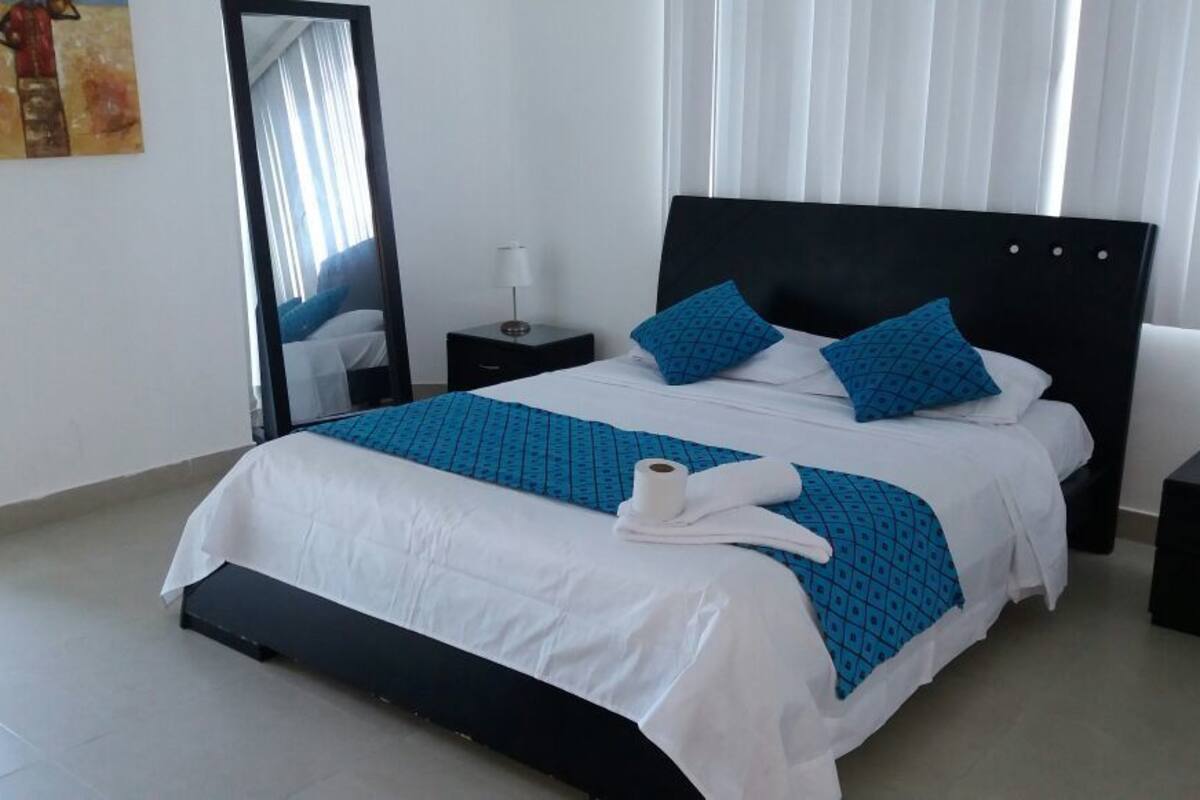 A neatly made bed with a white and blue bedspread, two pillows, and a rolled towel on a black headboard. There is a small bedside table with a lamp and a mirror on the wall.