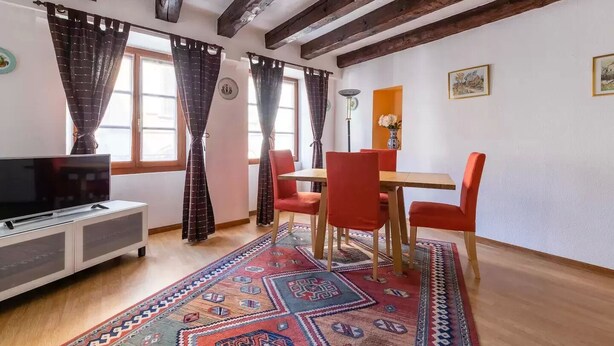 Een kamer met een televisie op een kast, een eettafel met rode stoelen en een kamerbreed tapijt met een patroon.