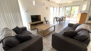 A modern living room with a flat-screen TV, a wooden coffee table, and two sofas with dark cushions. There is a dining area with a table and chairs visible through the glass doors. - Osprey Holiday Village Unit 111 - Villa with a Pool in the Complex (EXMOUTH)