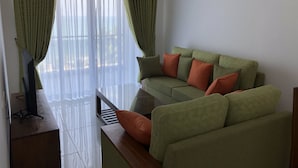 Un salon avec un canapé vert, des coussins orange, une table basse en verre et des rideaux légers.