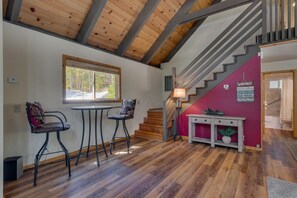 Een gezellige kamer met een houten plafond, een trap, een tafel met stoelen en een raam dat uitkijkt op groen.