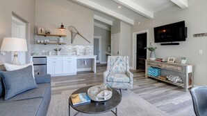 A spacious living room with a grey sofa, a coffee table with a book, a patterned armchair, a TV mounted on the wall, and a kitchen area with white cabinets and a hanging fishnet. - Ocean front, rock view: Salty Sister 3Bdrm with outdoor patios to gather (Morro Bay)