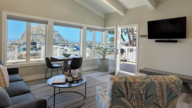 A spacious living room with large windows, a flat-screen TV, and a dining area with a table and chairs. - Ocean front, rock view: Salty Sister 3Bdrm with outdoor patios to gather (Morro Bay)